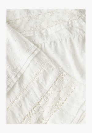 Biała tkanina z teksturowanymi kropkami, wstawkami z koronki i plisami z zakładek, tworząca delikatny, misterny wzór tekstylny.