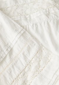 Tessuto di cotone bianco con delicata ricamo e dettagli in pizzo, con una superficie texturizzata e motivi sottili in tutto.