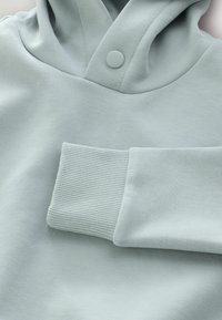 Jasnoniebieska bluza z kapturem o miękkiej, gładkiej fakturze, z żebrowanym mankietem i zapięciem na guziki przy kapturze. Prosty fason bez wzorów.