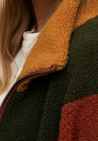 Fleecejakke med en orange krave, grøn krop og røde detaljer, med lynlås og blød tekstur.