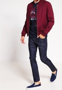 Burgundy bomberjacka med dragkedja framtill, marinblå grafisk t-shirt, mörka jeans och blå slip-on skor med bruna accenter.