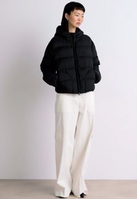Melna pūkā vestē ar kapuci, ar rāvējslēdzēju priekšā un elastīgām piedurknēm, kombinēta ar platām baltām biksēm ar sānu kabatām.