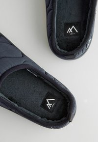 Pantofole in tessuto trapuntato blu navy con rivestimento soffice, design slip-on e suola texturizzata. Logo visibile sulla soletta. Aspetto morbido e accogliente.