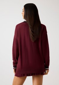 Maglione lavorato a maglia colore mogano con maniche lunghe, polsini a coste con strisce blu navy e bianche, e orlo plissettato, visto da dietro.