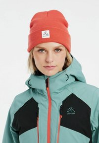 Berretto arancione a coste e giacca zip con colori teal, nero e arancione, caratterizzata da un logo prominente e un design geometrico. Texture liscia, dettagli funzionali.