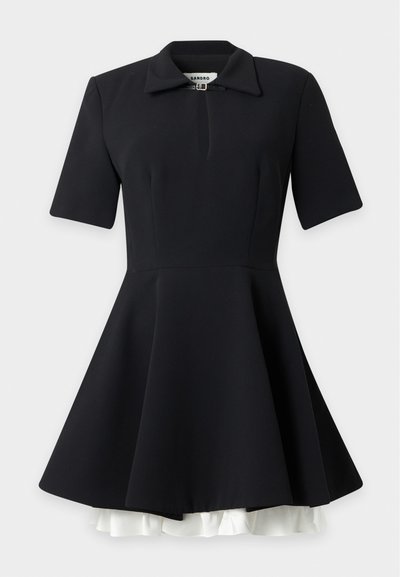 Robe noire à manches courtes avec une jupe évasée, col, petit détail de boucle au niveau de l'encolure et bordure à volants blanche dépassant de l'ourlet.