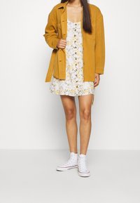 Žlutá džínová bunda přeložená přes bílou květinovou šaty s žlutými a šedými akcenty. Bílé vysoké tenisky dotahují celý outfit.