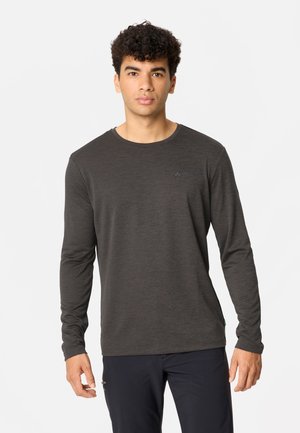 Junger Mann mit lockigem Haar, der ein dunkelgraues Langarmshirt und dunkle Hosen trägt, steht vor einem schlichten weißen Hintergrund.