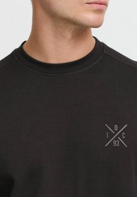 Black sweatshirt with a textured finish, featuring a round neck and a small graphic in the lower left corner with the letters "IDC 93."