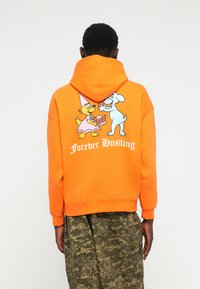 Felpa arancione con cappuccio che presenta personaggi dei cartoni animati sul retro, con la scritta "Forever Hustling" in bianco. Il materiale sembra morbido e testurizzato.