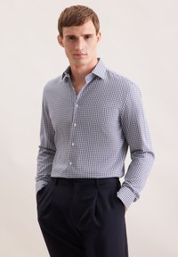 Giovane uomo che indossa una camicia a scacchi blu e bianca con bottoni infilata in pantaloni a pieghe blu navy scuri, con le mani in tasca.