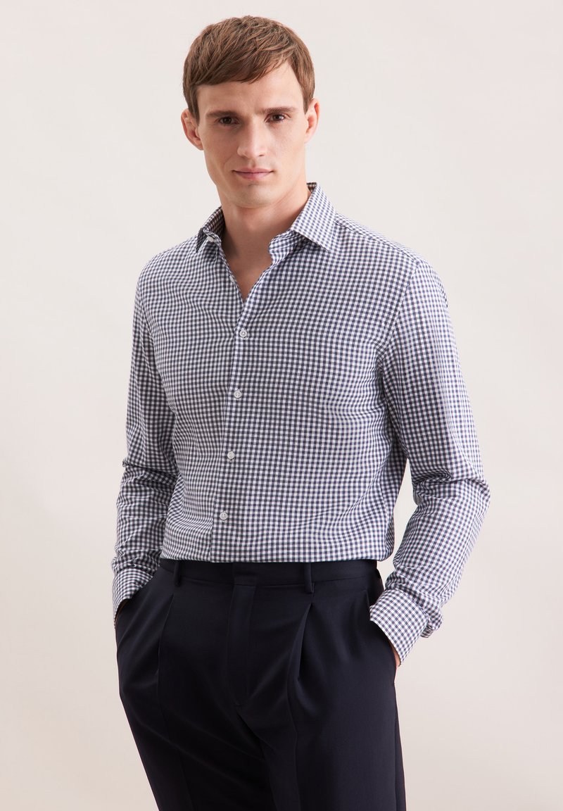 Giovane uomo che indossa una camicia a scacchi blu e bianca con bottoni infilata in pantaloni a pieghe blu navy scuri, con le mani in tasca.