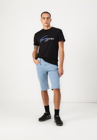 Svart bomull t-skjorte med hvit og blå "Tommy Jeans" logo, kombinert med lyseblå denimshorts. Svart joggesko med sokker.