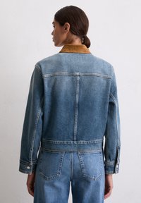 Veste en denim bleu clair avec un col en daim marron. Comprend des poignets à boutons et des détails sur le dos, coupée dans une silhouette cropped.