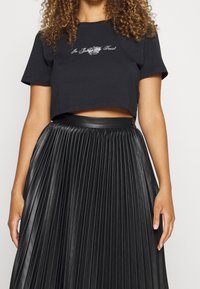 T-shirt noir court à manches courtes avec broderie blanche ; associé à une jupe plissée noire taille haute en matériau brillant.