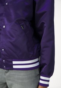 Paarse satijnen bomberjack met drukknoopsluiting, zijzakken en witte gestreepte ribgebreide mouwen. Beschikt over een klein merklabel op de zak.