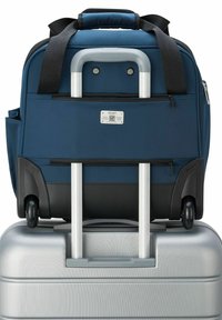 Sac à dos en tissu bleu marine avec une poignée rétractable, posé sur une valise argentée texturée. Présente des accents noirs et des roues pour la mobilité.