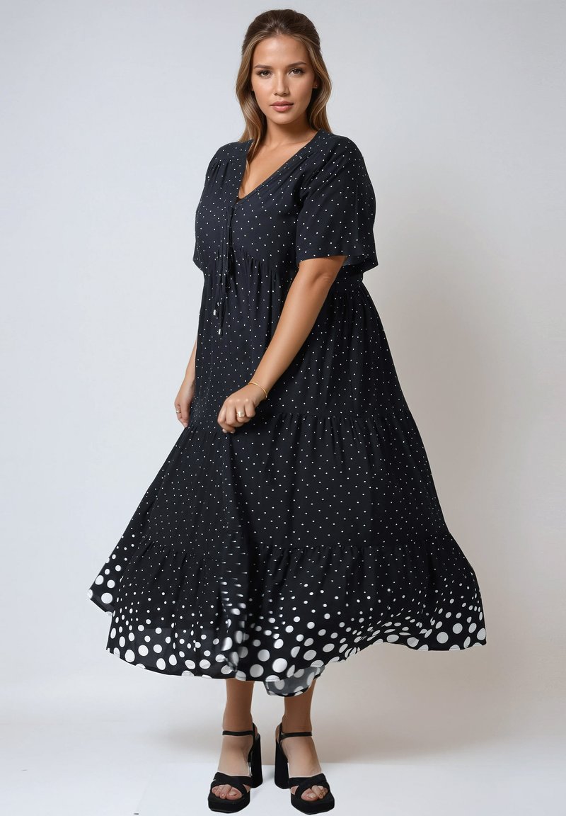 Zwarte maxi-jurk met stippen, korte mouwen, v-hals, verzameld middenstuk en een gelaagde zoom, met grotere witte stippen onderaan.