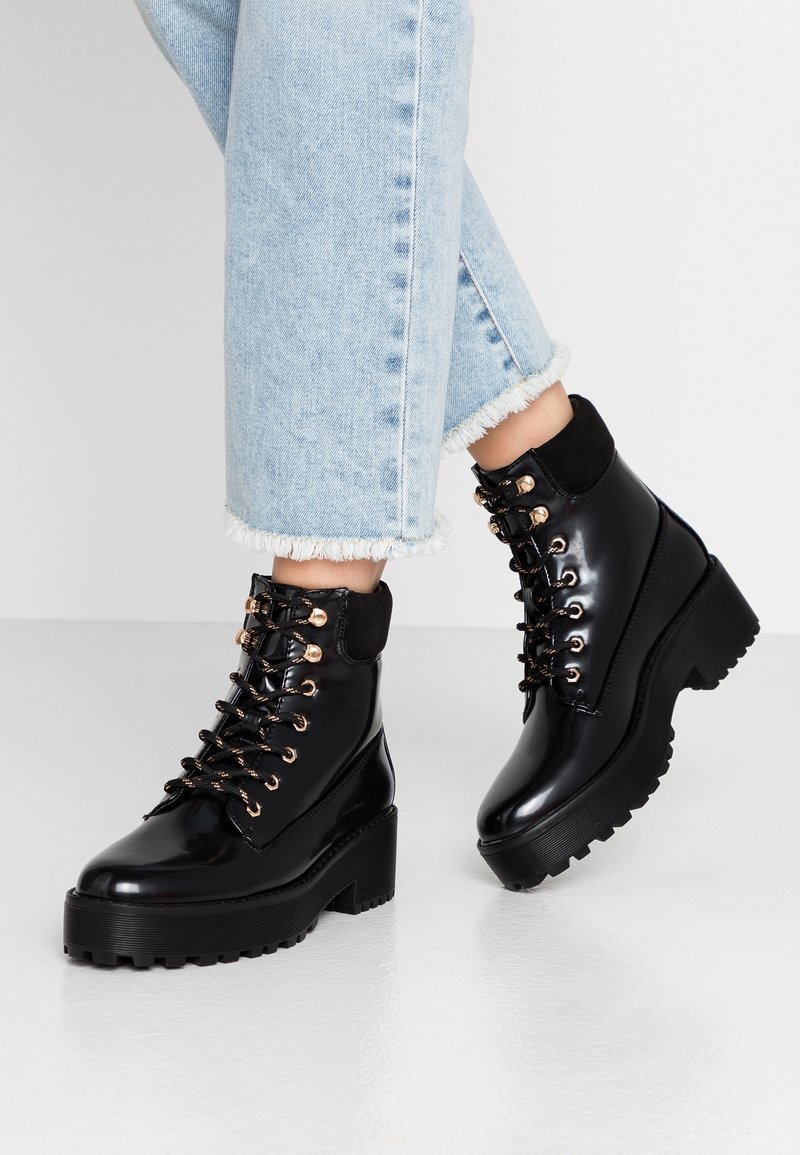 Bottes de cheville noires brillantes avec une semelle chunky noire, une quincaillerie de lacet dorée et un col en daim. Associées à un jean bleu clair effiloché.