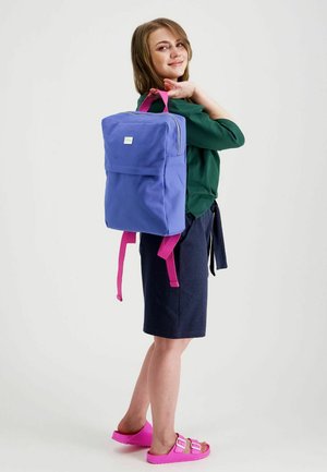 Nuori nainen, joka pukeutuu vihreään toppiin, tummansiniseen hameeseen ja vaaleanpunaisiin sandaaleihin, kantaa olkapäällään sinistä reppua, jossa on vaaleanpunaiset remmit.