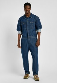 Combinaison en denim bleu foncé avec des manches longues, deux poches poitrine et une taille cintrée. Assortie à des baskets multicolores.