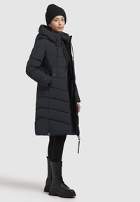 Doudoune marine avec capuche amovible, design matelassé, poches latérales et fermeture éclair. Assortie à des bottes de combat noires à lacets.