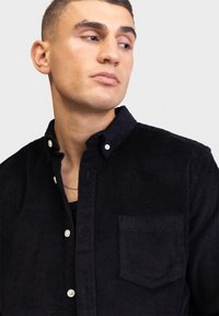Camicia in corduroy nero con colletto button-down, tasca frontale e bottoni bianchi. La texture è a coste, aggiungendo profondità al tessuto.