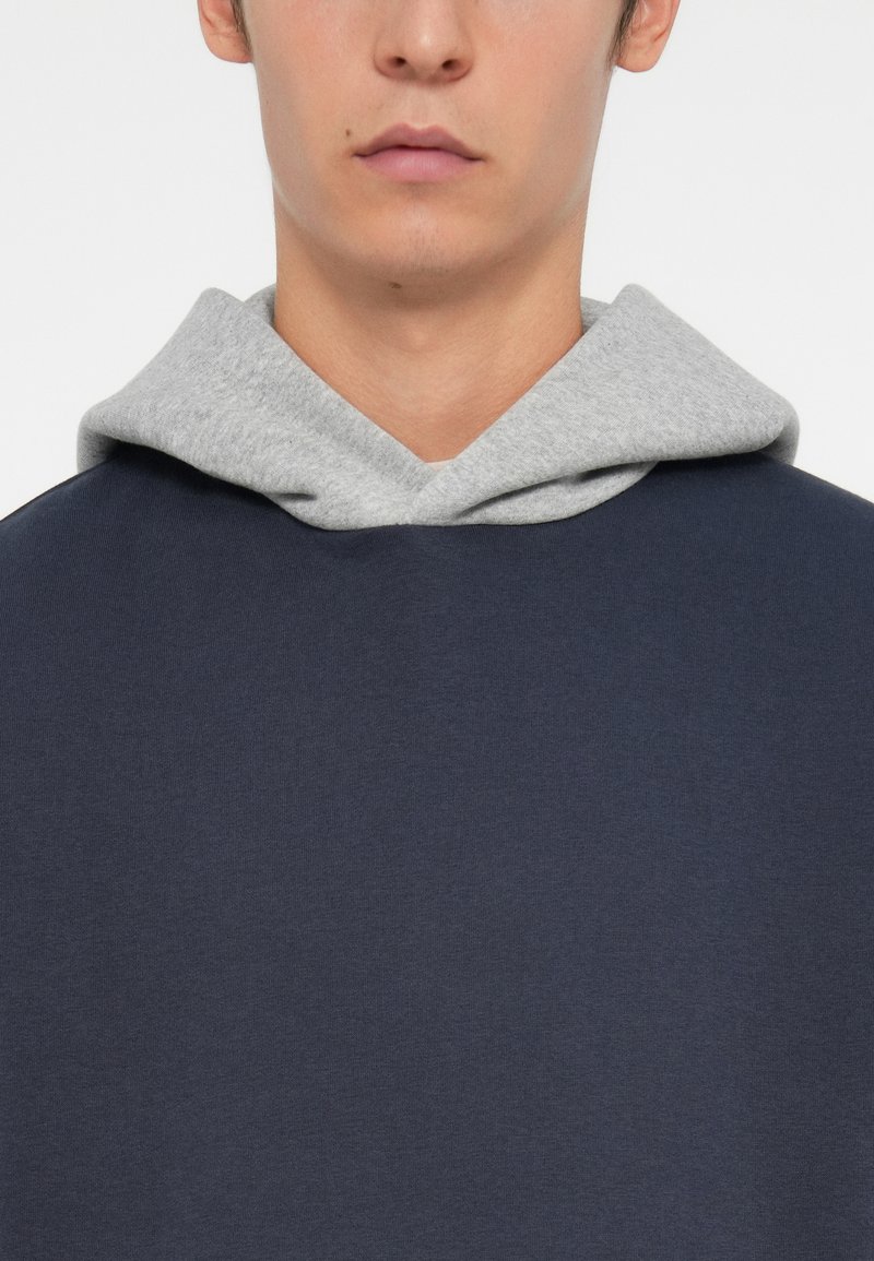 Marineblauer Pullover mit grauem Kapuzen; weicher Baumwollmischstoff; lockere Passform; minimale Nähte; keine sichtbaren Logos oder Verzierungen.