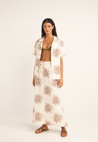 Conjunto ligero de dos piezas de crema que incluye una camisa con botones y una falda maxi con patrones de sol marrones. Combinado con un bikini verde y sandalias.