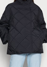 Zwarte gewatteerde jas met een hoge kraag, voorzien van een rechte silhouet en diamantpatronen. Draagt over lichtblauwe spijkerbroeken.