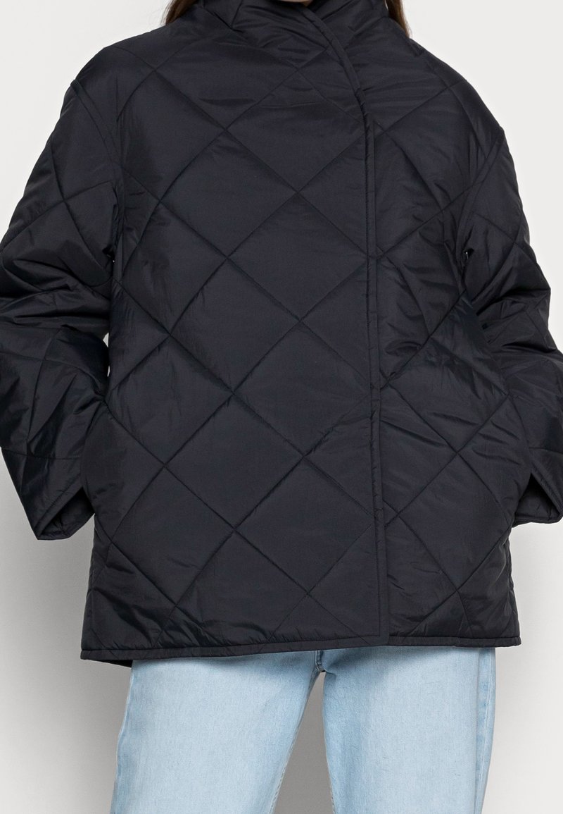 Zwarte gewatteerde jas met een hoge kraag, voorzien van een rechte silhouet en diamantpatronen. Draagt over lichtblauwe spijkerbroeken.