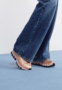 Sportieve sneakers met een glanzende roségouden bovenkant, golvend ontwerp en zwarte rubberen zool. Gecombineerd met wijde, donkerdenim jeans op een blauwe vloer.