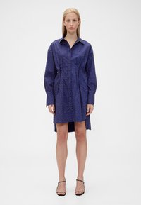 Blue button-up dress with a pointed collar, long sleeves, elastic waist, and white polka dot pattern, featuring a high-low hem.