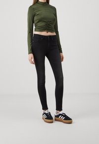 Svarta skinny jeans tillsammans med en kortärmad, olivgrön topp med insynad detaljer och svarta sneakers med vita ränder.