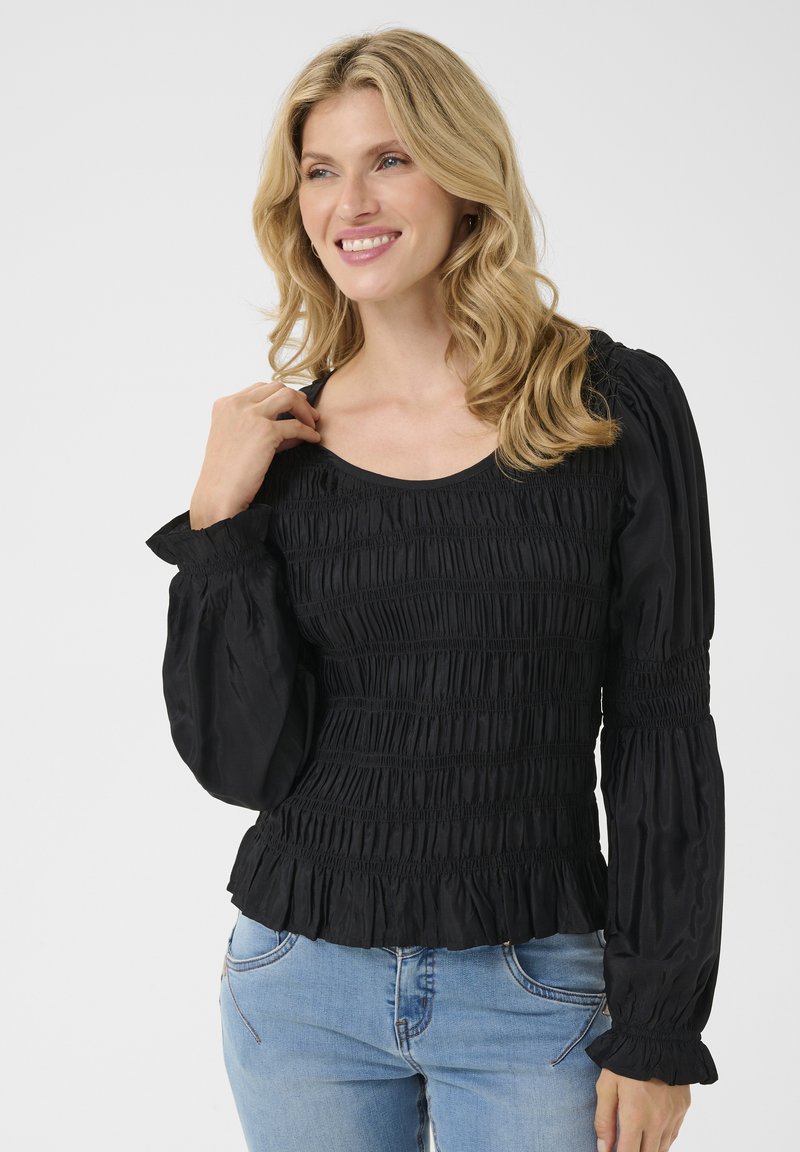 Haut smocké noir avec de longues manches bouffantes et un ourlet à volants. Tissu doux avec une texture froncée sur toute la surface. Associé à un jean bleu clair.