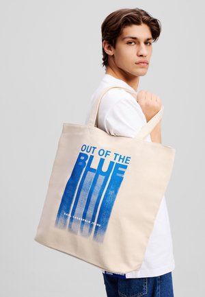 Canvas tote bag i lys beige med blå grafisk tekst som leser "OUT OF THE BLUE." Har solide håndtak og jevn tekstur.