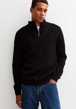 Pull en maille noir avec un col zippé quart et une texture côtelée, associé à un jean en denim bleu. Le pull a une coupe décontractée.
