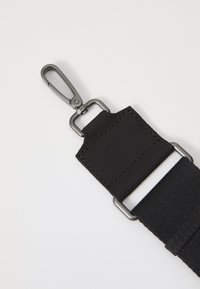 Sangle noire avec un attachement en métal. Présente un tissu texturé et un patch en cuir lisse avec des détails de couture. Design minimaliste.