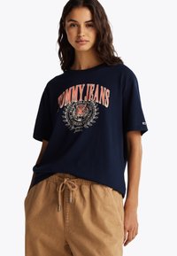 Marinblå bomulls T-skjorte med et stort tigerdesign og "TOMMY JEANS"-tekst i korall, kombinert med beige bukser med snøring.