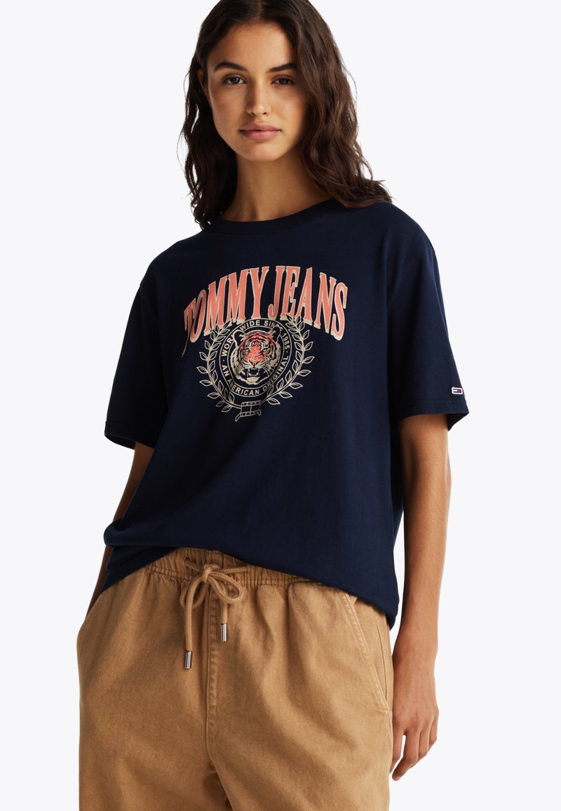 Marinblå bomulls T-skjorte med et stort tigerdesign og "TOMMY JEANS"-tekst i korall, kombinert med beige bukser med snøring.