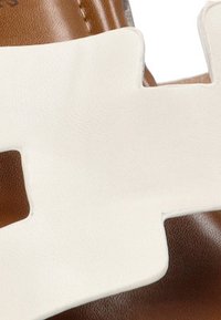 Sandal de cuero marrón con un diseño de tira recortada en blanco. La tira es suave con una textura brillante, proporcionando contraste con el marrón.