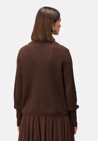 Maglione marrone a coste con scollatura alta e maniche lunghe, caratterizzato da delicate decorazioni. Indossato sopra una gonna a pieghe marrone coordinata.