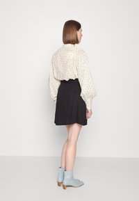 Blusa creme com padrões florais, mangas balão e saia preta. A modelo usa botas a ankle azul claras e posa de costas.