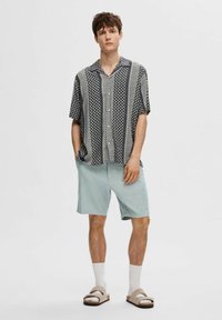 Camicetta a maniche corte con motivo geometrico blu navy e bianco, abbinata a pantaloni corti verde chiaro, calzini bianchi e sandali beige, su uno sfondo chiaro.