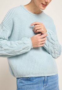 Hellblau gestrickter Pullover mit strukturiertem Körper, dicken Zopfmuster-Ärmeln und rundem Halsausschnitt. Kombiniert mit hellblauen Jeans.