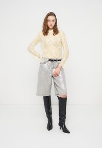 Haut à manches longues en dentelle jaune clair, assorti à un short argenté brillant et à des bottes noires montantes à bout pointu.