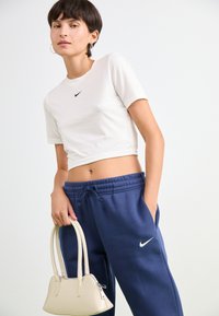 Ungdom med kort hår som har på seg en hvit cropped T-skjorte og blå Nike joggebukser, holder en kremfarget håndveske med glidelås.