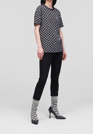 Camisa corta de manga corta con estampado en blanco y negro, jeans negros ajustados y tacones altos negros con calcetines texturizados y estampados, de pie contra un fondo liso.