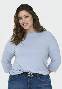 Lys blå langermet topp med avslappet passform og båtutringning, parret med et svart belte med dobbelt sirkulært spenne, iført denimjeans.