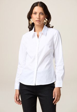 Camisa blanca de manga larga y botón hecha de algodón. Presenta un cuello clásico y un ajuste entallado. Se lleva con pantalones negros adornados con pequeños remaches.